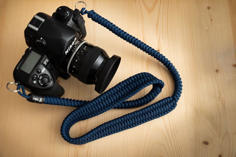 May include: A black DSLR camera with a lens attached, sitting on a wooden surface. The camera is connected to a dark blue braided camera strap with silver metal rings.