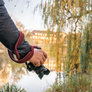 May include: A person holding a black camera with a red camera strap in front of a body of water and trees.