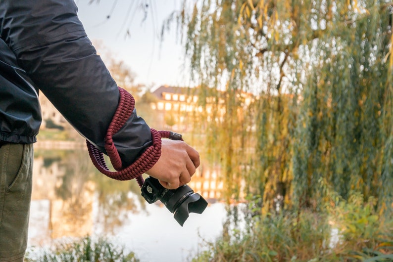 May include: A person holding a black camera with a red camera strap in front of a body of water and trees.