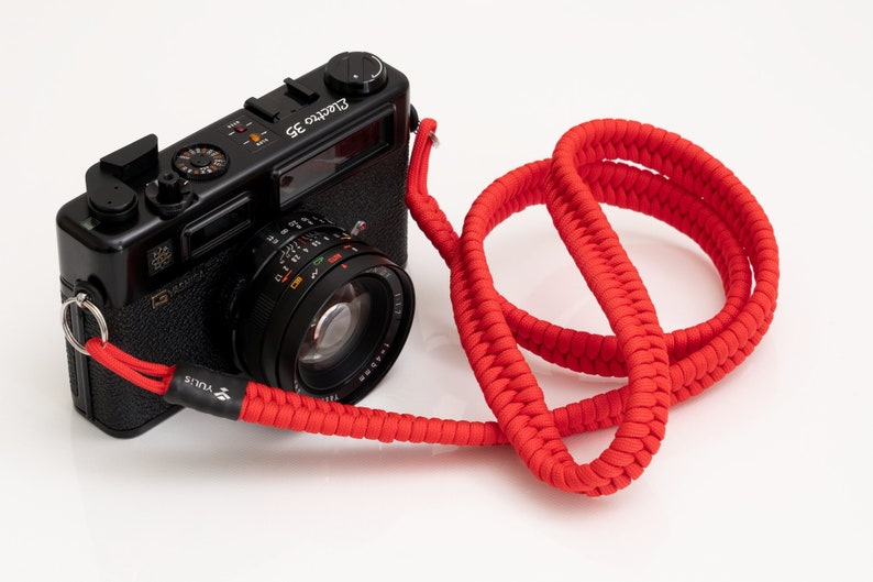 May include: A black 35mm film camera with a red braided camera strap. The camera has a lens with a silver ring and the text "Electra 35" on the top.
