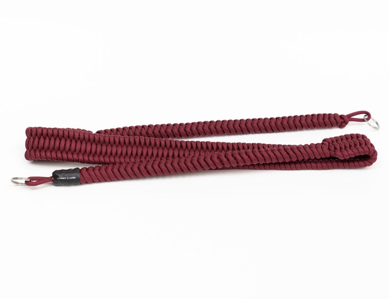 May include: A maroon braided camera strap with metal rings at each end.