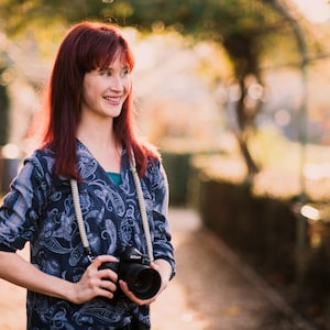 May include: A woman with red hair is holding a black camera with a white strap. She is wearing a blue and white patterned shirt and is standing in a park with trees and green foliage in the background.