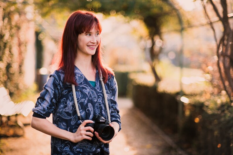 May include: A woman with red hair is holding a black camera with a white strap. She is wearing a blue and white patterned shirt and is standing in a park with trees and green foliage in the background.