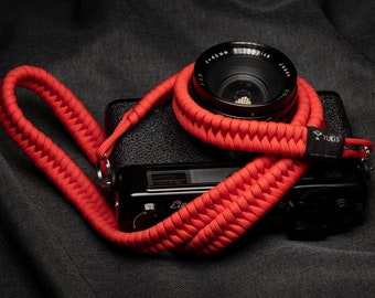 May include: A red camera strap made of braided paracord with a black camera attached. The strap has a Yulis logo on it.