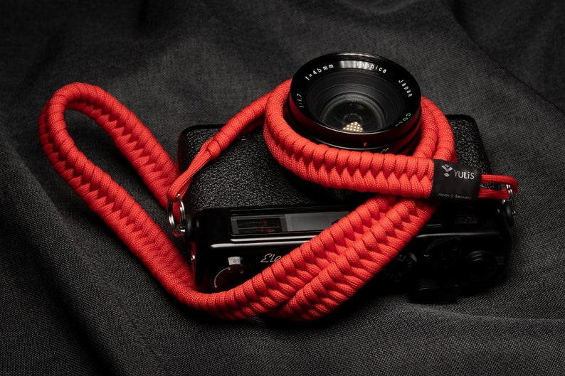 May include: A red camera strap made of braided paracord with a black camera attached. The strap has a Yulis logo on it.