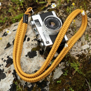 May include: A mustard yellow braided camera strap with a silver camera attached. The camera is a YASHICA Electro 35 with a TELE-YASHINON 35mm lens. The camera and strap are lying on a rock covered in moss and small white flowers.
