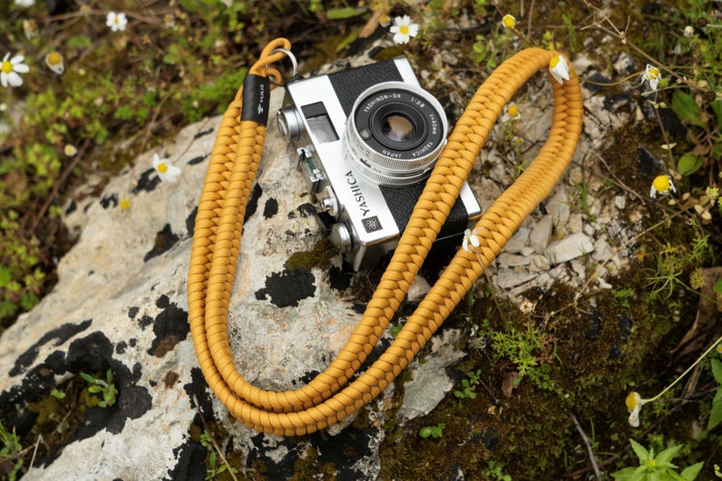 May include: A mustard yellow braided camera strap with a silver camera attached. The camera is a YASHICA Electro 35 with a TELE-YASHINON 35mm lens. The camera and strap are lying on a rock covered in moss and small white flowers.