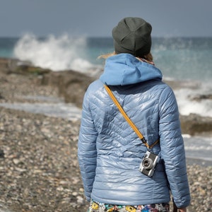 May include: A person wearing a blue hooded jacket and a dark gray beanie stands on a rocky beach, looking out at the ocean waves. A camera is attached to a yellow strap around their neck.
