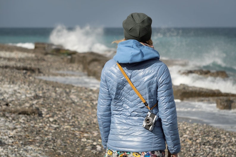 May include: A person wearing a blue hooded jacket and a dark gray beanie stands on a rocky beach, looking out at the ocean waves. A camera is attached to a yellow strap around their neck.