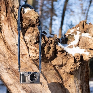 May include: A silver camera with a blue braided strap hanging from a tree branch covered in snow.