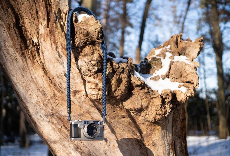 May include: A silver camera with a blue braided strap hanging from a tree branch covered in snow.