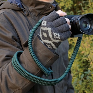 May include: A dark green camera strap made of braided cord. The strap is worn around the wrist and attached to a black camera.