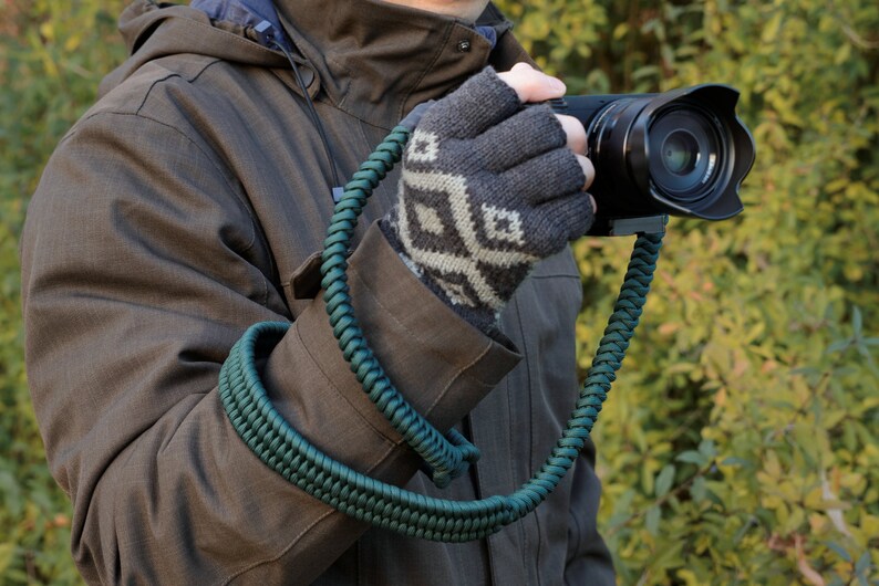 May include: A dark green camera strap made of braided cord. The strap is worn around the wrist and attached to a black camera.