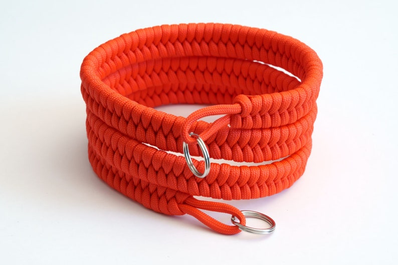 May include: A bright orange paracord bracelet with two silver key rings. The bracelet is made with a woven pattern.