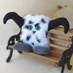 Puede incluir: Un juguete de peluche blanco y negro con grandes ojos verdes se sienta en un pequeño banco de madera con patas de metal negro.