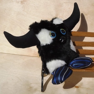 Puede incluir: Juguete de peluche negro y blanco con ojos azules y cuernos. El juguete lleva pantalones a rayas azules y blancas y está sentado en un banco de madera.