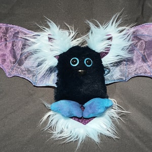 May include: A black plush bat with blue wings and white fur. The bat has large blue eyes and a small black nose.