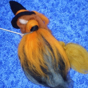 May include: A handmade felted witch doll with a black hat and orange hair. The doll has a wooden stick and a yellow broom. The witch is made of wool and sits on a blue patterned background.