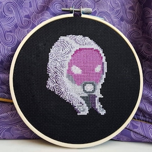May include: A cross-stitch embroidery hoop with a black background and a white and pink pixelated image of a hooded figure.