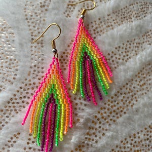 May include: A pair of vibrant, beaded earrings in a teardrop shape. The earrings feature a gradient of neon colors, including pink, orange, yellow, green, and purple. Gold-colored ear wires complete the design, creating a bold and colorful accessory.