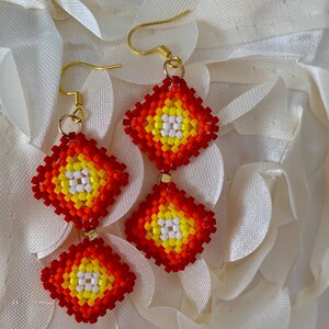 May include: Handmade beaded earrings with a geometric design. The earrings feature a diamond shape with a vibrant color palette of red, yellow, and white beads. Gold-colored ear wires complete the design. The earrings are displayed on a white, textured surface.