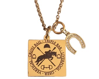 May include: Gold-coloured necklace with a square pendant depicting a horse and rider jumping, surrounded by text. A horseshoe charm, embellished with clear stones, dangles beside the pendant. The chain is also gold-coloured.