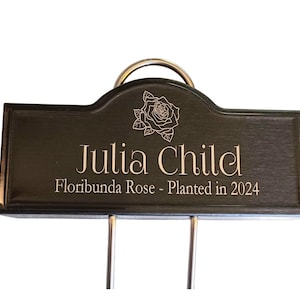 Op de afbeelding: Een donkerbruine houten tuinmarker met een metalen ring aan de bovenkant. De marker is gegraveerd met de tekst "Julia Child" en "Floribunda Rose - Geplant in 2024".