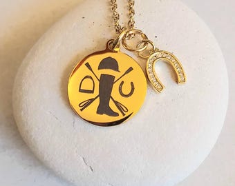 May include: Gold necklace with a round pendant featuring a silhouette of a rider with crossed riding crops and the letters "D" and "C". A small horseshoe charm with clear stones is attached to the pendant.