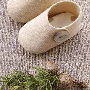 Op de afbeelding: Een paar crèmekleurige vilten pantoffels met een knoopsluiting. De pantoffels zijn gemaakt van een zacht, getextureerd materiaal en hebben een ronde neus. De knoop is parelmoer. De pantoffels liggen op een getextureerd oppervlak.