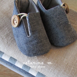 Puede incluir: Un par de zapatos de bebé de mezclilla azul con un botón de madera en un zapato. Los zapatos están sobre una tela de lino beige.