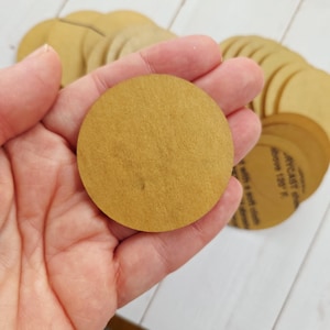 May include: A stack of circular, gold-colored acrylic discs with a matte finish. Each disc is flat and uniform in shape. One disc is held in the foreground, while several others are layered behind it, showing partial text on the bottom disc.