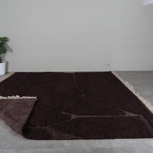 May include: A large, rectangular brown rug with a shaggy texture. The rug has a cream-colored fringe along the edges. Part of the rug is folded over, revealing the underside. The rug is placed on a gray floor.
