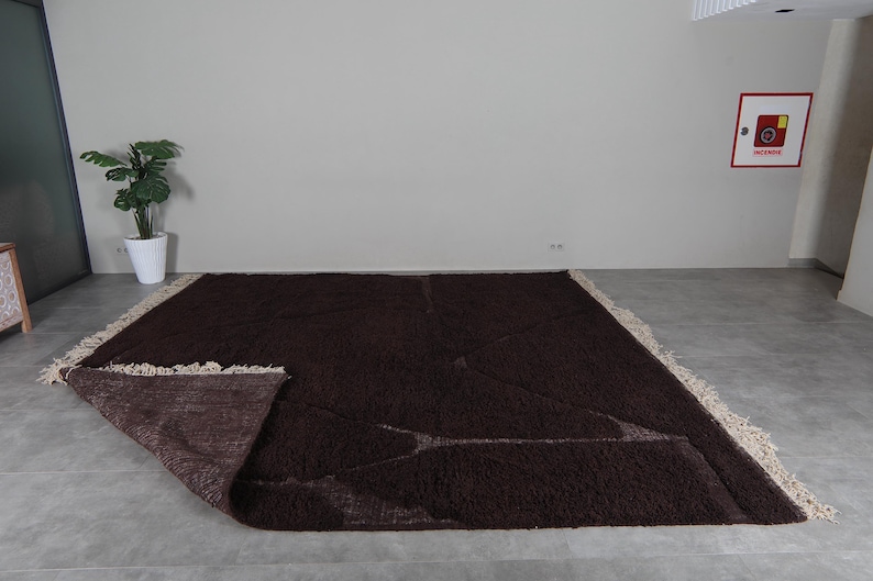 May include: A large, rectangular brown rug with a shaggy texture. The rug has a cream-colored fringe along the edges. Part of the rug is folded over, revealing the underside. The rug is placed on a gray floor.