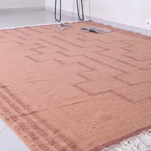 May include: A light brown woven rug with a geometric pattern in a darker brown shade. The rug has a fringe border.