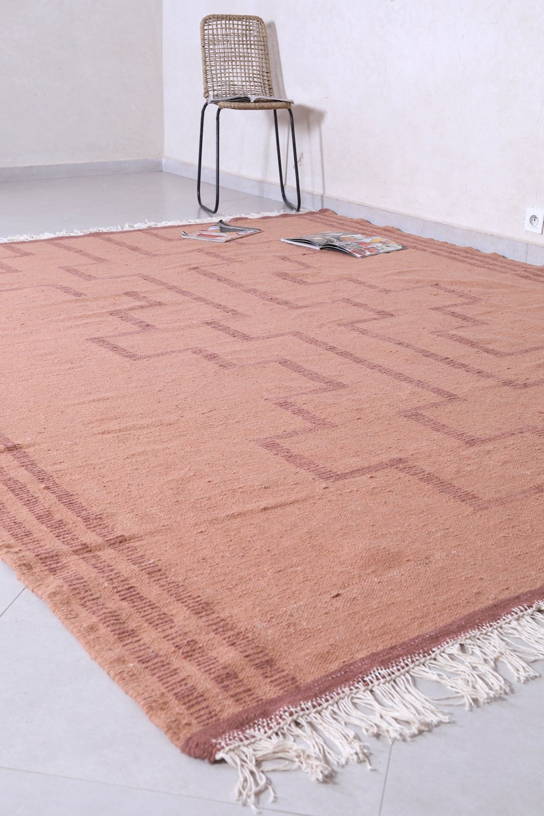 May include: A light brown woven rug with a geometric pattern in a darker brown shade. The rug has a fringe border.