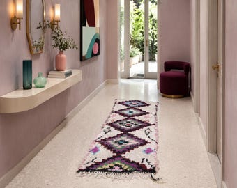 Può includere: Un lungo tappeto da corridoio rettangolare con base color crema e motivi a rombi ripetuti in viola scuro, nero e verde acqua. Il tappeto presenta una consistenza a pelo lungo con estremità sfrangiate ed è posizionato in un corridoio.