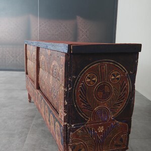 May include: An antique wooden chest with hand-painted designs in brown, red, and blue. The chest has a rectangular shape with a flat top and is supported by four short legs. The artwork includes circular and floral motifs. The chest is likely made of wood.