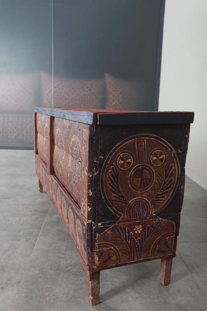 May include: An antique wooden chest with hand-painted designs in brown, red, and blue. The chest has a rectangular shape with a flat top and is supported by four short legs. The artwork includes circular and floral motifs. The chest is likely made of wood.