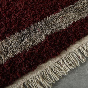 May include: Close-up of a textured rug with a burgundy and gray striped pattern. The rug has a cream-colored fringe along the edge. The rug's design is a mix of burgundy and gray, creating a visually appealing texture.