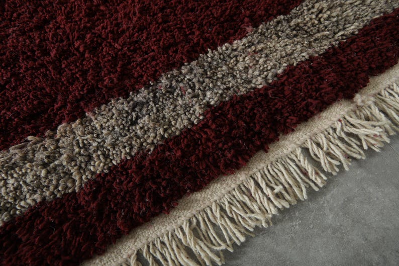 May include: Close-up of a textured rug with a burgundy and gray striped pattern. The rug has a cream-colored fringe along the edge. The rug's design is a mix of burgundy and gray, creating a visually appealing texture.