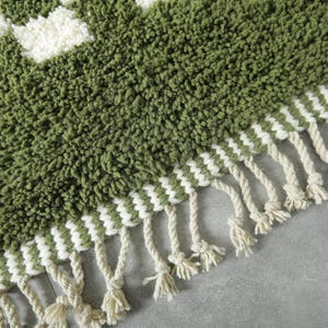 May include: Close-up of a green and white patterned rug with a textured, shaggy surface. The rug features a geometric design with white squares and a fringe of cream-colored tassels along the edge. The rug is made of a soft, plush material.