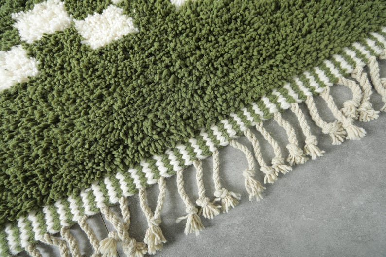 May include: Close-up of a green and white patterned rug with a textured, shaggy surface. The rug features a geometric design with white squares and a fringe of cream-colored tassels along the edge. The rug is made of a soft, plush material.