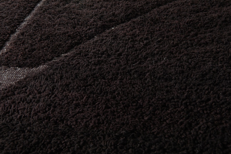 May include: Close-up of a dark brown, textured fabric. The material has a dense, looped pile, creating a soft, plush appearance. A lighter, textured area is visible, possibly a seam or a different fabric section.