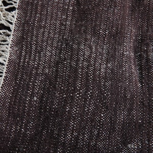 May include: Close-up of a dark brown rug with a textured weave and off-white fringe. The rug's surface has a speckled appearance due to the lighter-colored threads woven into the darker base. The fringe is thick and made of a cream-colored yarn.