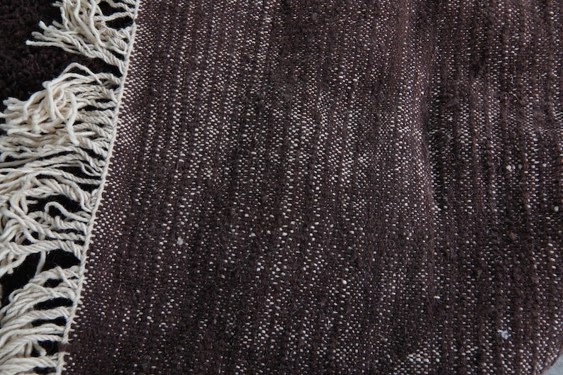 May include: Close-up of a dark brown rug with a textured weave and off-white fringe. The rug's surface has a speckled appearance due to the lighter-colored threads woven into the darker base. The fringe is thick and made of a cream-colored yarn.