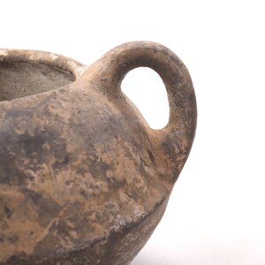May include: A brown clay pot with a single handle. The pot has a rough, textured surface and appears to be very old.