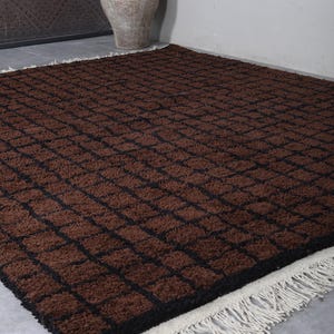 Peut inclure: Un tapis rectangulaire marron avec un motif quadrill&eacute; de lignes marron fonc&eacute;. Le tapis a une bordure noire et une frange cr&egrave;me le long d'un bord. Le tapis est pos&eacute; sur un sol gris.
