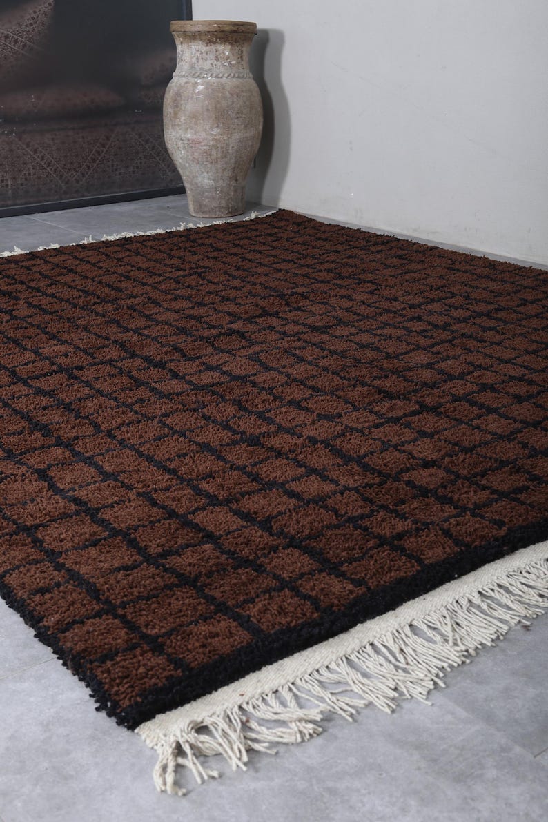 Peut inclure: Un tapis rectangulaire marron avec un motif quadrill&eacute; de lignes marron fonc&eacute;. Le tapis a une bordure noire et une frange cr&egrave;me le long d'un bord. Le tapis est pos&eacute; sur un sol gris.