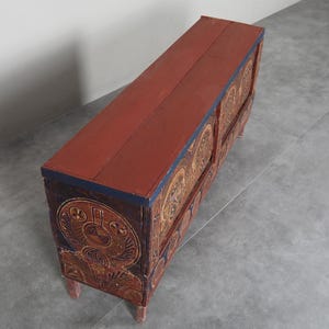 May include: A long, rectangular wooden chest with a red top and intricately carved sides. The sides feature a repeating pattern of circular designs in gold and brown. The chest has four short legs and a dark blue trim.