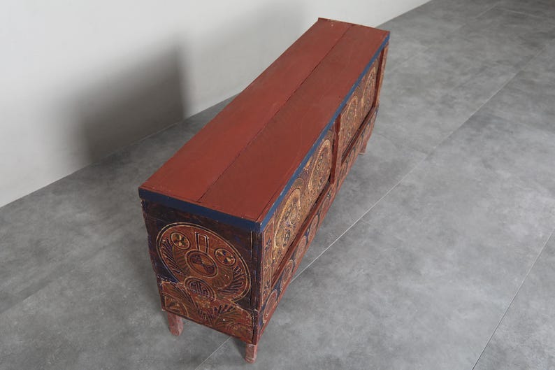 May include: A long, rectangular wooden chest with a red top and intricately carved sides. The sides feature a repeating pattern of circular designs in gold and brown. The chest has four short legs and a dark blue trim.